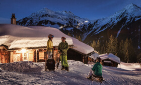 Zwei Personen stehen neben ihren Rodel und eine Person sitzt startbereit auf dem Rodel auf der Schneepiste