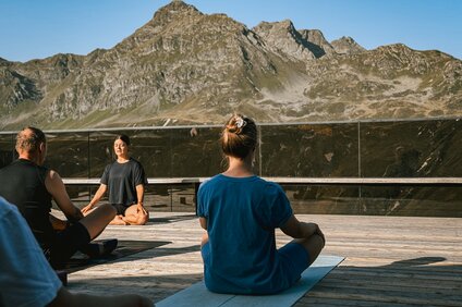 Eine Gruppe von Personen sitzt auf einer Terrasse bei Morgensonne und macht Yoga. | © Silvretta Montafon - Vanessa Strauch