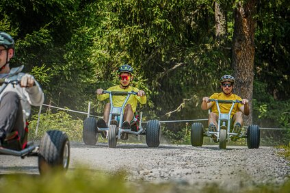 Drei junge Männer fahren auf Mountaincarts einen Güterweg im Wald entlang. | © Silvretta Montafon - Vanessa Strauch