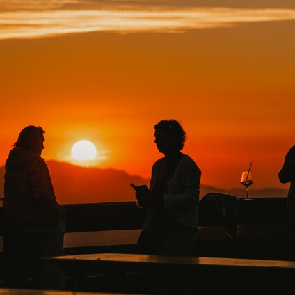 3 Personen die den Sonnenuntergang im Kapellrestaurant am Geländer in der Silvretta Montafon genießen | © Silvretta Montafon - Vanessa Strauch