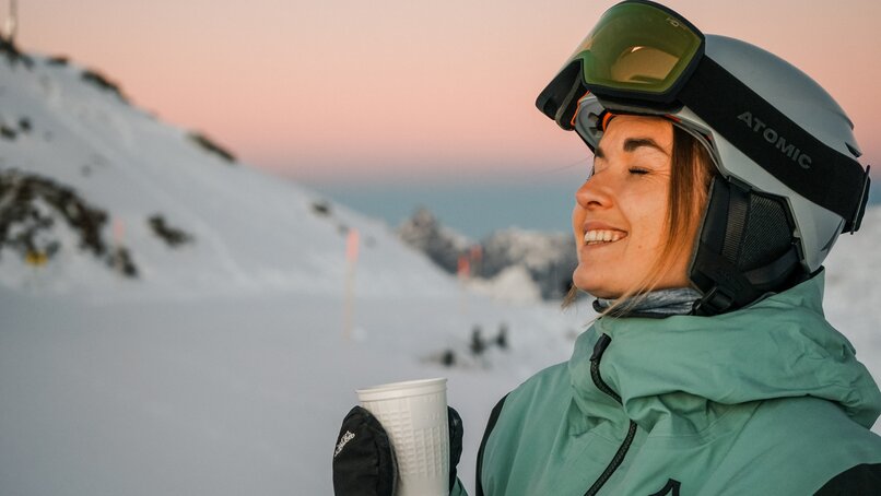 Eine Frau mit Skibrille und Skihelm genießt einen Tee bei Sonnenaufgang am Berg. | © Silvretta Montafon - Vanessa Strauch