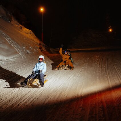 Drei Personen in Skikliedung rodeln auf einem Schlitten am Abend. | © Silvretta Montafon - Marie Schilcher