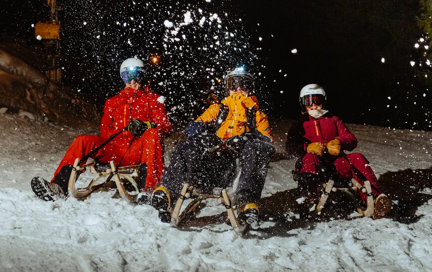 Drei Personen rodeln auf einem Schlitten und der Schnee spritzt in die Kamera. | © Silvretta Montafon - Marie Schilcher