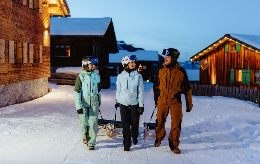Drei Personen in Skikleidung ziehen einen Rodel durch ein verschneites Bergdorf am Abend. | © Silvretta Montafon - Marie Schilcher