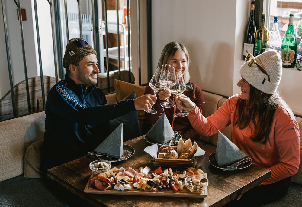 Drei Personen an einem Tisch mit Käse und Brot sitzen in einer Weinstube und stoßen mit einem Glas Weißwein an. | © Silvretta Montafon - Vanessa Strauch