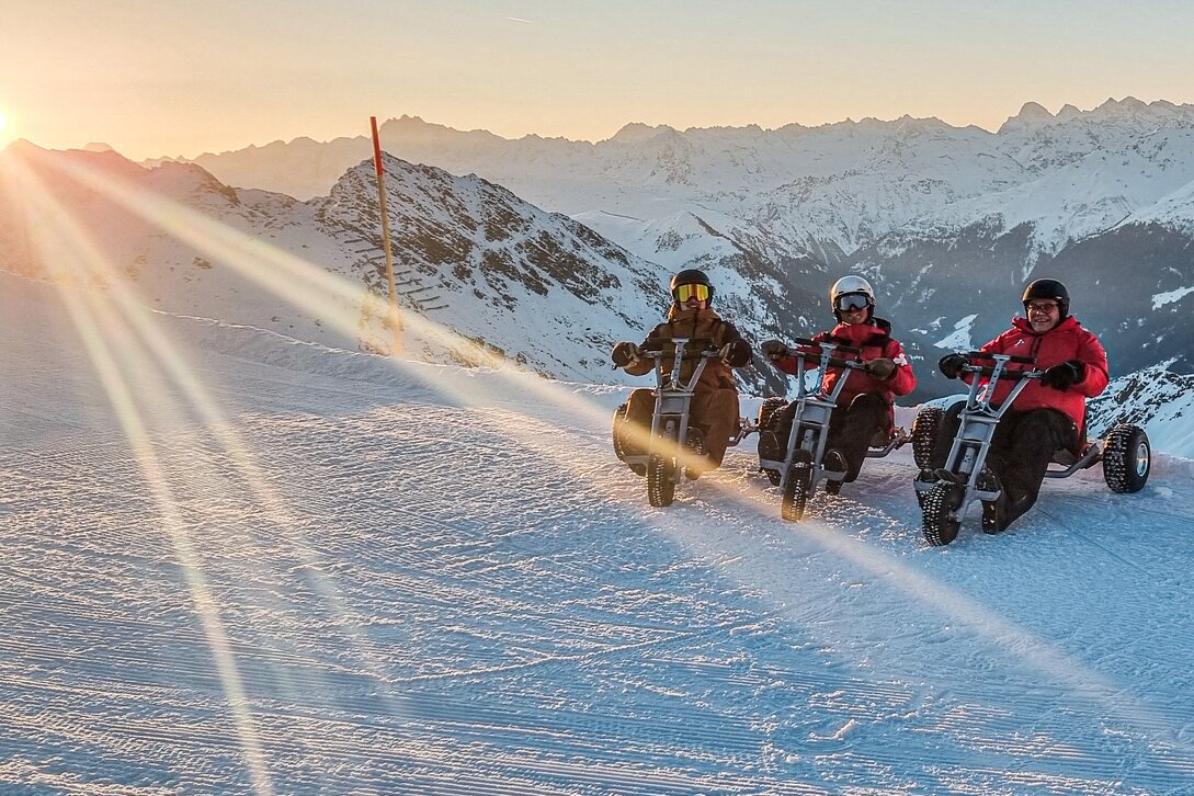Drei Männer sitzen auf einem Mountain Cart im Schnee bei Sonnenaufgang auf der Skipiste. | © Silvretta Montafon