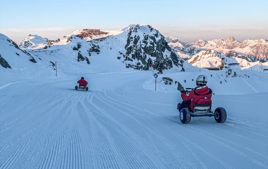 Zwei Personen fahren auf einem Mountain Cart im Schnee bei Sonnenaufgang auf der Skipiste. | © Silvretta Montafon
