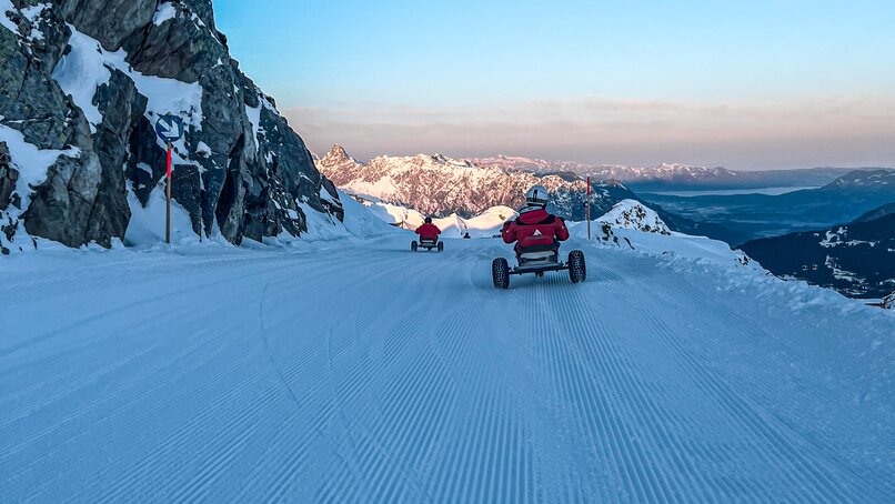 Zwei Personen fahren auf einem Mountain Cart im Schnee bei Sonnenaufgang auf der Skipiste. | © Silvretta Montafon