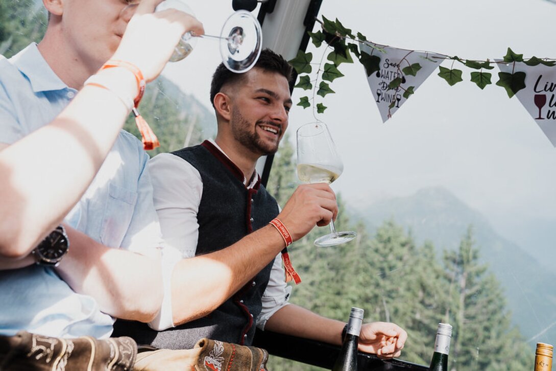 Ein junger Mann sitzt in Tracht in einer Gondel und genießt ein Glas Weißwein | © Silvretta Montafon - GOODGUYPAEZ