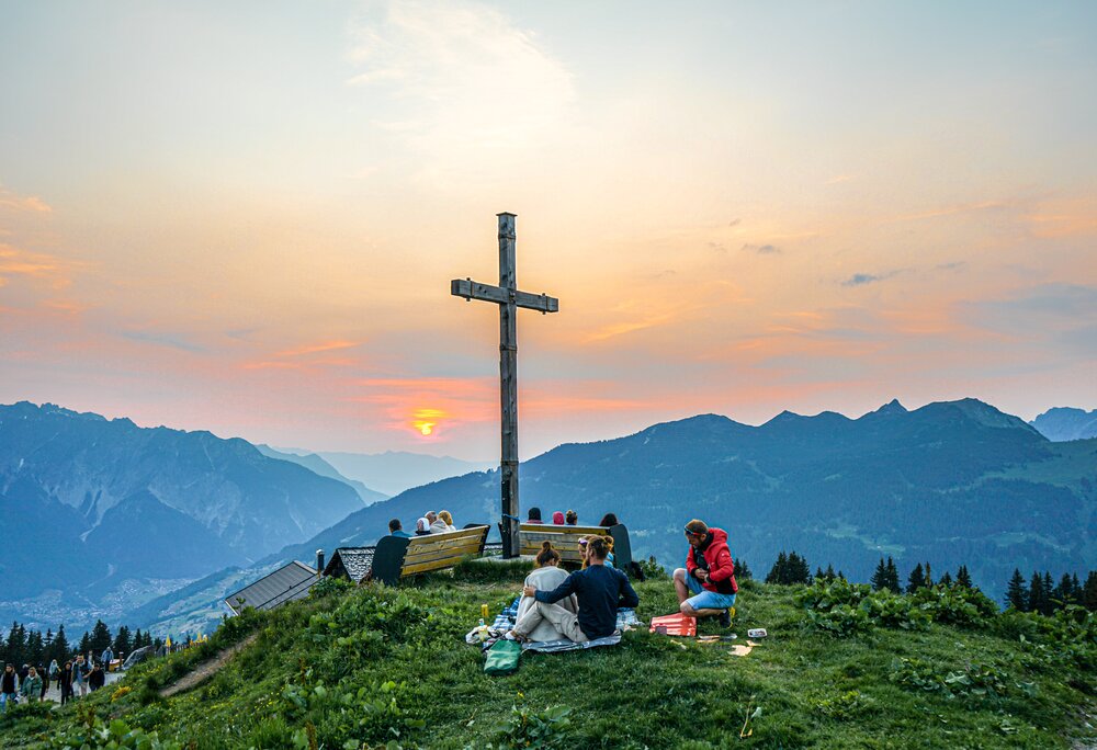 Viele Personen stehen am Gipfelkreuz und genießen den Sonnenuntergang bei der Sonnwendfeier in der Silvretta Montafon. | © Silvretta Montafon - Vanessa Strauch
