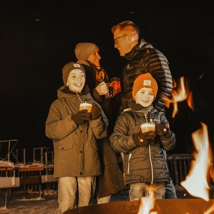 Eine Familie mit zwei Kindern steht mit einem warmen Getränk am Feuer vor einer Berghütte. | © Vanessa Strauch - Silvretta Montafon