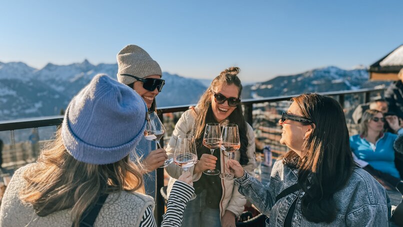 Vier junge Frauen stoßen mit einem Glas Wein an bei Sonnenaufgang auf einer Terrasse im Skigebiet. | © Vanessa Strauch - Silvretta MOntafon