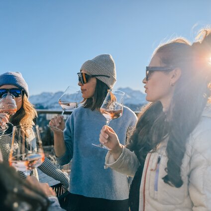Drei junge Frauen mit einem Glas Wein bei Sonnenaufgang auf einer Terrasse im Skigebiet. | © Silvretta Montafon - Vanessa Strauch